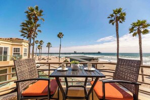 A balcony with wicker chairs and orange cushions, overlooking a beach with palm trees and a pier. - 5N-64 Beachfront On the Strand (Oceanside)