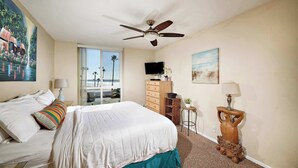A hotel room with a bed, a ceiling fan, a TV, a dresser, and a view of the beach through the window. - North Coast Village D204 (Oceanside)