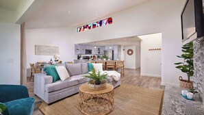 Ein modernes Wohnzimmer mit einem grauen Sofa, einem blauen Sessel und einem Korbtisch. Im Hintergrund befindet sich eine Küche mit grauen Schränken und ein Essbereich mit Korbstühlen.