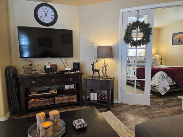 A living room with a flat-screen TV, a wooden side table with a lamp, a fireplace, and a bed with a Christmas wreath.