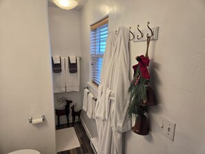 A bathroom with white walls, a wooden floor, a window with blinds, a towel rack with white towels, a decorative pine branch, and a toilet paper holder.