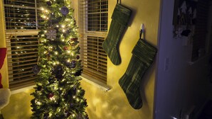 Un sapin de Noël décoré d’ornements et de guirlandes lumineuses, une fenêtre avec des stores et une chaussette verte accrochée au mur.