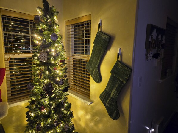 A Christmas tree with ornaments and lights, a window with blinds, and a green stocking hanging on the wall.