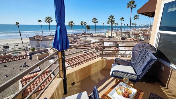 Un balcón con vistas a la playa, palmeras y un área de comedor con mesa y sillas.