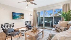 A modern living room with a ceiling fan, a large TV, wicker chairs, a sofa, and a view of palm trees through the glass door. - Sunrise & Sunset by the Sea  812-1 (Oceanside)