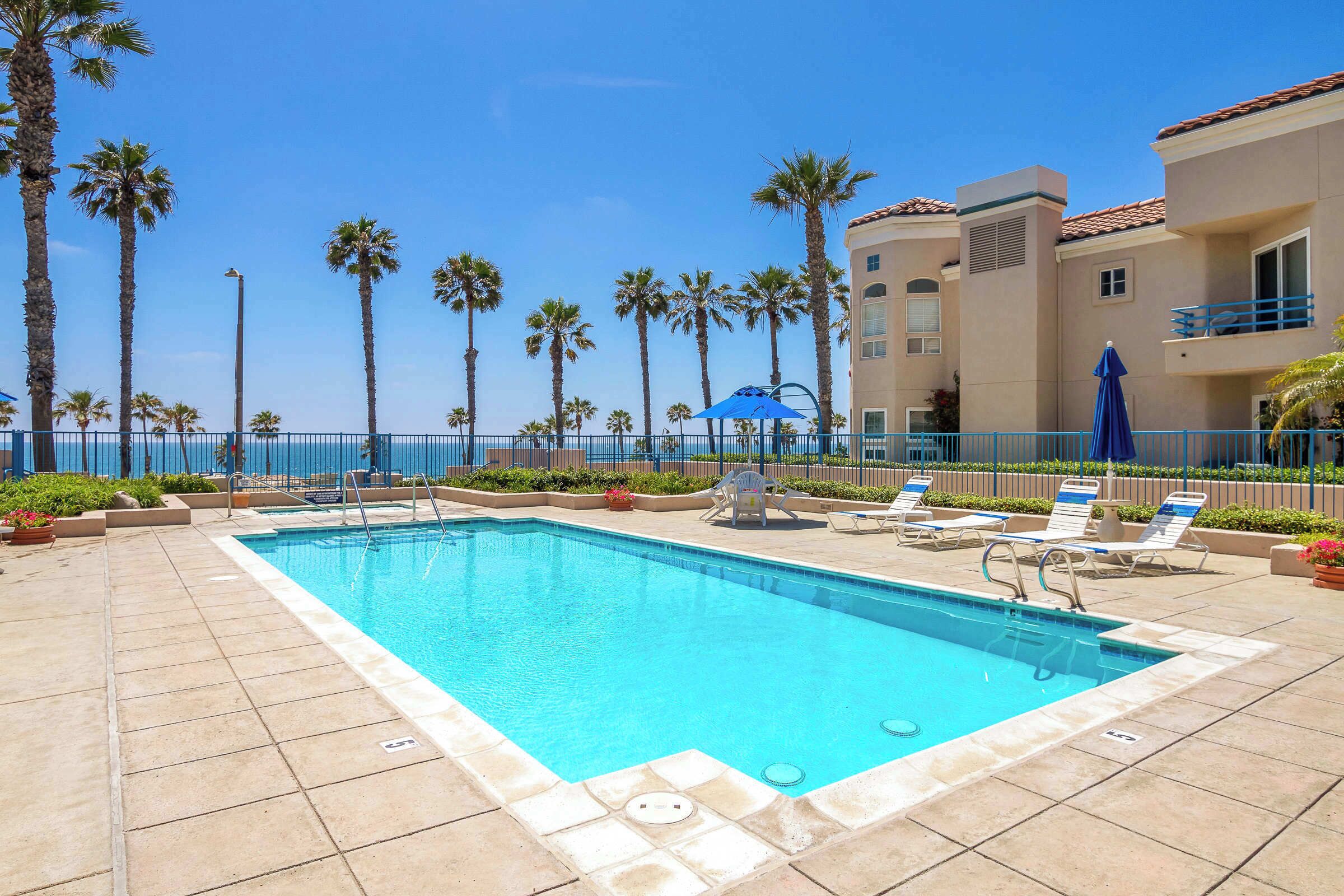 A swimming pool with lounge chairs and umbrellas, surrounded by palm trees and a building with a balcony.