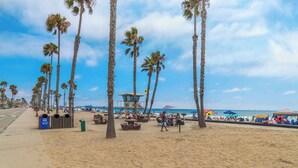 Une plage bordée de palmiers, avec un poste de surveillance pour les sauveteurs, des tables de pique-nique et des parasols.