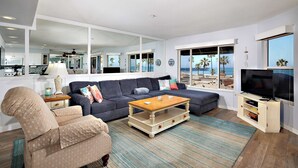 Un salon spacieux avec un grand canapé, une table basse et un téléviseur. Les fenêtres offrent une vue sur la plage.