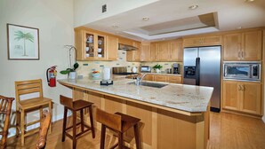 A modern kitchen with wooden cabinets, a stainless steel refrigerator, a microwave, and a large island with bar stools. - 152-4 Coastal Luxury with Panoramic Ocean View (Oceanside)
