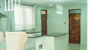 A kitchen with a white countertop, a sink, and a window with curtains. There are two wooden doors and a white cabinet.