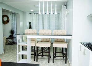 Un coin repas comprenant une table et quatre chaises, un ventilateur, un espace cuisine avec des placards et un plancher de couleur claire.