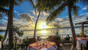 A deck with tables set for dining, overlooking a body of water with a sunset. - Hôtel KOU-BUGNY (L'Ile-des-Pins)