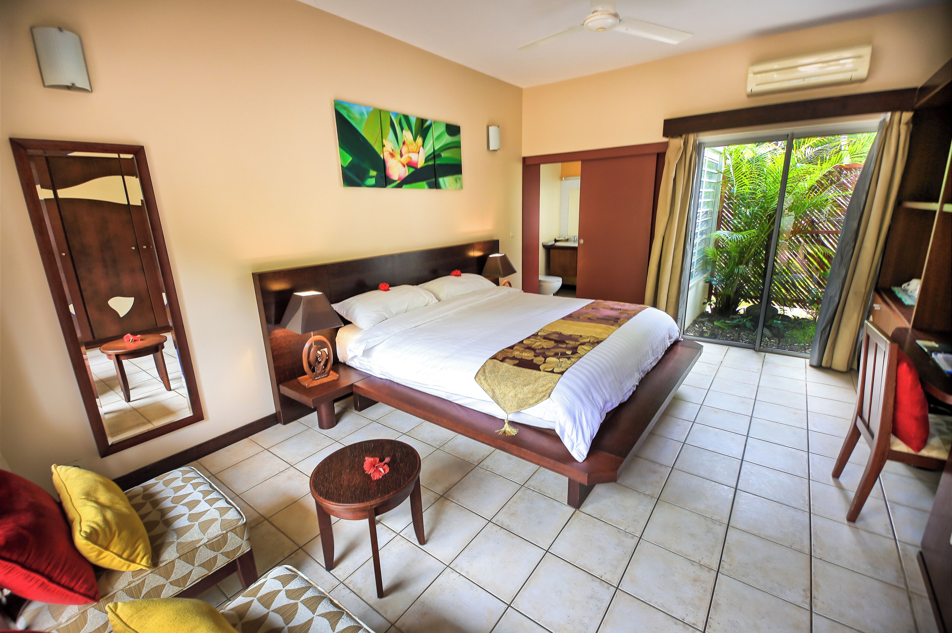 A hotel room with a large bed, a bedside table, a small round table, a chair, and a large window with a view of greenery.