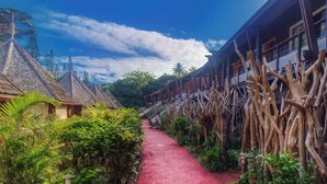 A pathway flanked by traditional wooden buildings with thatched roofs, surrounded by lush greenery and a clear blue sky. - Hôtel KOU-BUGNY (L'Ile-des-Pins)