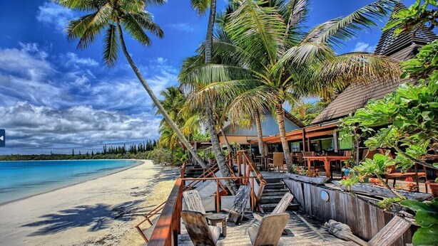 A wooden deck with lounge chairs and a restaurant area with outdoor seating, overlooking a beach with palm trees and a clear blue sky. - Hôtel KOU-BUGNY (L'Ile-des-Pins)
