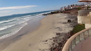 A beach with a rocky shoreline, a pier in the background, and a building with an umbrella. - Oceanfront Rosarito Balcony, Beach & Amazing Sunsets (Playas de Rosarito)