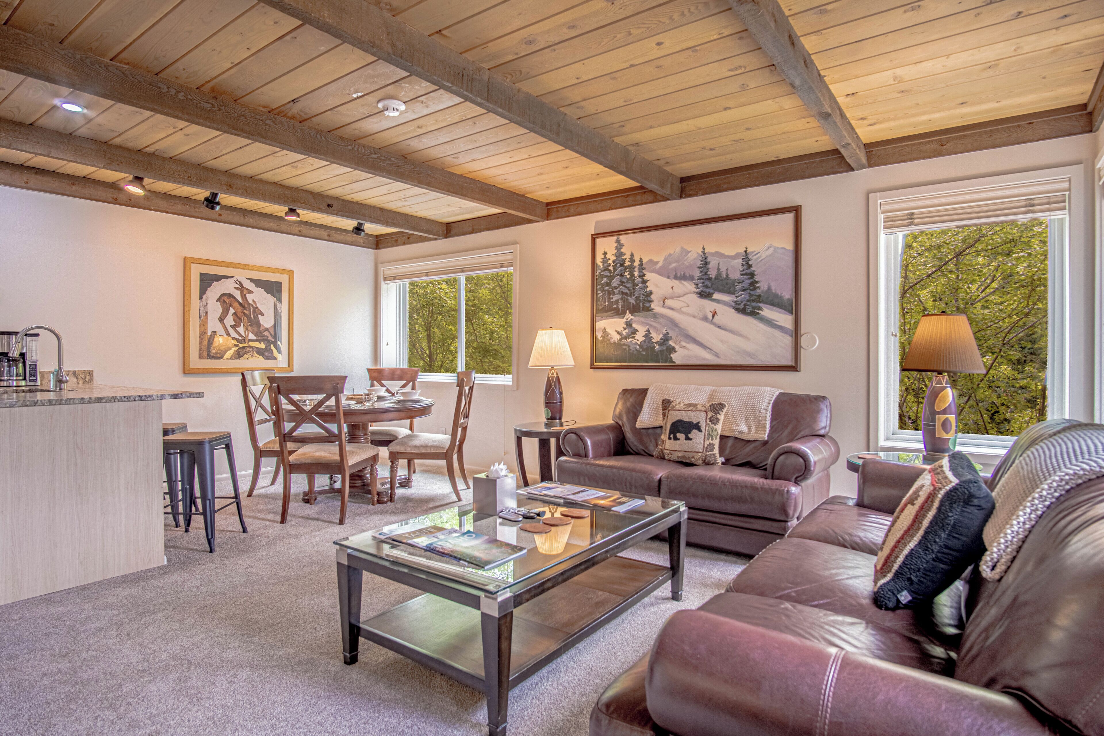 Un salon chaleureux avec un plafond en bois, un coin repas et une grande fenêtre donnant sur les arbres.