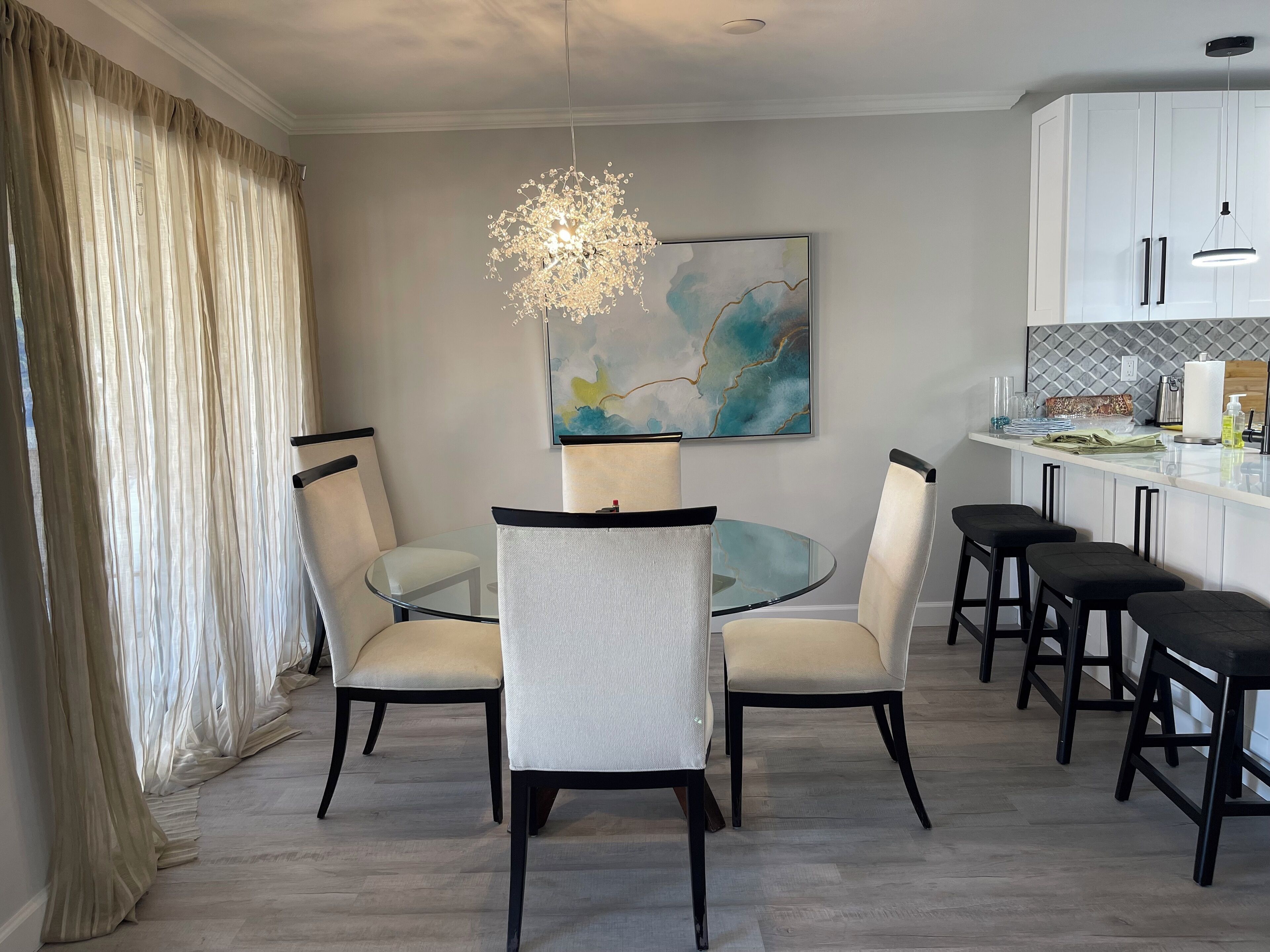 Un espace de salle à manger moderne, avec une table en verre entourée de chaises, un lustre et une œuvre d’art contemporaine accrochée au mur.