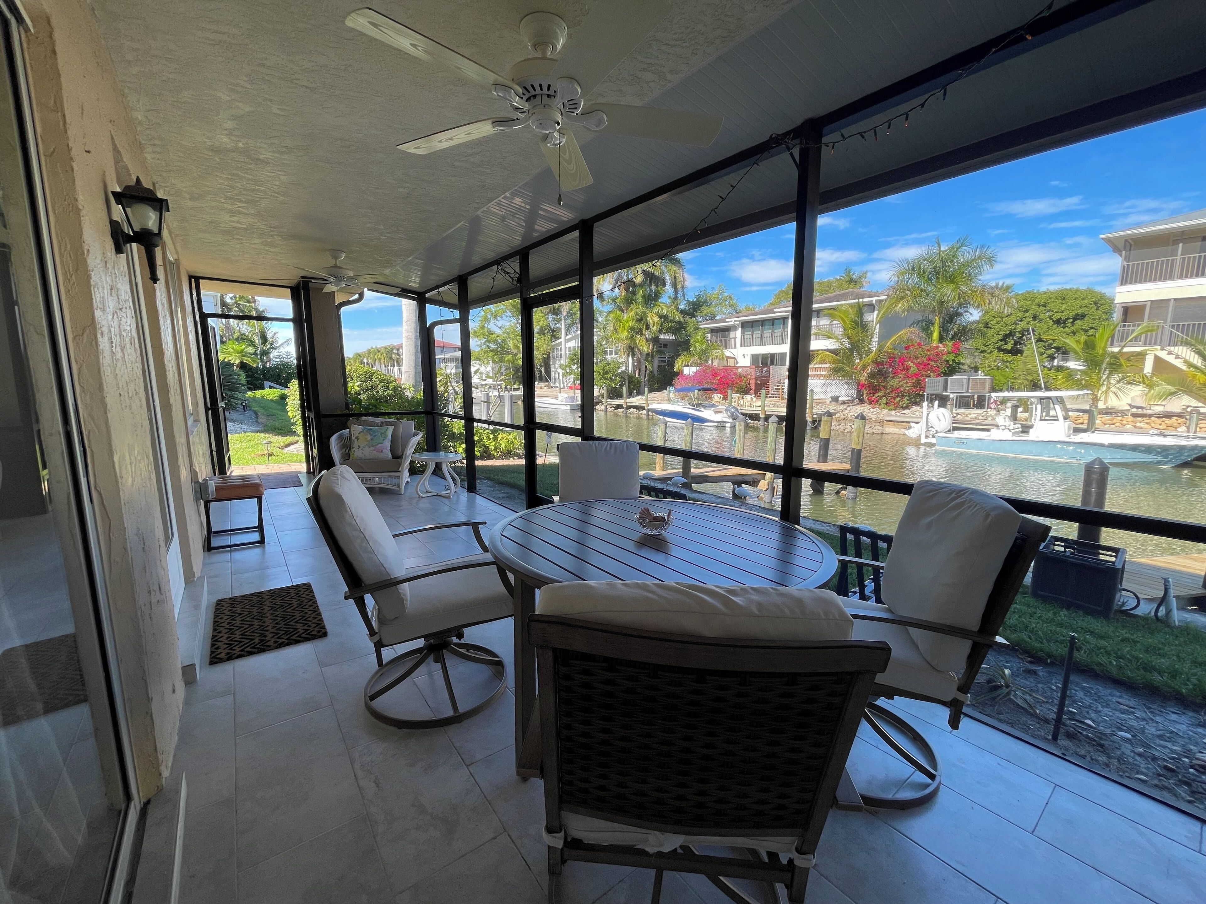 A covered outdoor seating area with a table and chairs, large windows offering a view of a pool and buildings, and a ceiling fan.