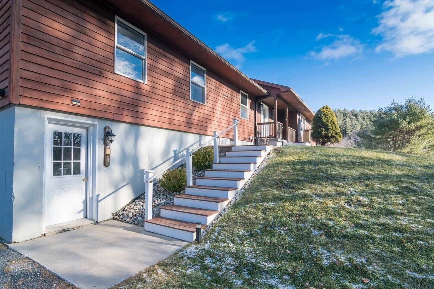 Ett tvåvåningshus med en veranda framtill, en trappa som leder upp till ytterdörren och en liten trädgård med gräsmatta och några träd.
