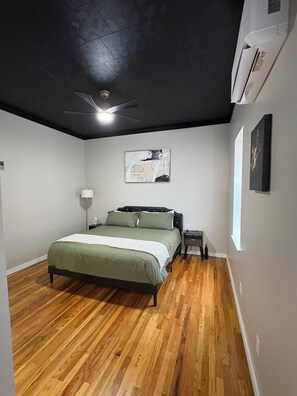 Une chambre à coucher comprenant un lit, un ventilateur de plafond, une table de chevet et un climatiseur fixé au mur.
