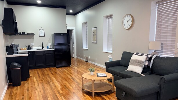 A modern living room with a dark grey sofa, a small wooden coffee table, and a kitchen area with black cabinets and a black refrigerator.