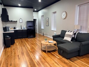 Una sala de estar moderna con un sofá gris oscuro, una pequeña mesa de centro de madera y una cocina con gabinetes negros y un refrigerador negro.