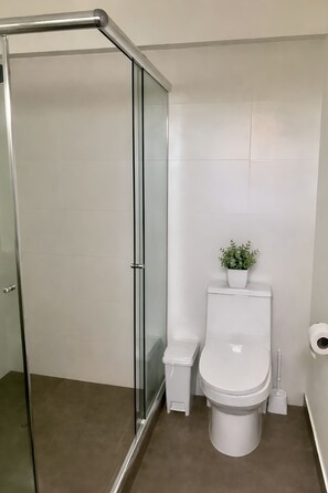 A modern bathroom with a glass shower enclosure, a white toilet, a small trash bin, and a potted plant on the toilet tank. - Modern 2-Bedroom Apartment in Miraflores | King-Size Bed & Terrace with Park Views (Miraflores)