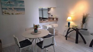 Ein Essbereich mit rundem Tisch, weißen Stühlen, einer Vase mit Blumen, einem kleinen Küchentisch mit Lampe und einem Bild von einer Strandlandschaft.