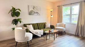 Una sala de estar con un sofá verde, dos sillones blancos, una mesa de centro de vidrio, una lámpara de pie, una planta en maceta y una ventana con cortinas.
