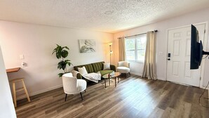 Una sala de estar con un sofá verde, sillones blancos, una mesa de centro, una lámpara de pie, un cuadro en la pared y una puerta con un televisor montado.