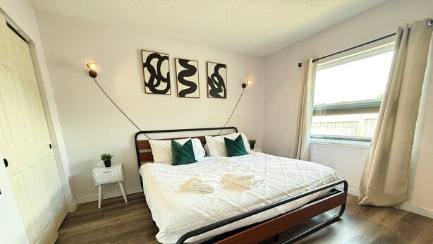 Un dormitorio con cama, almohadas, una mesita de noche con una planta y una obra de arte en la pared.