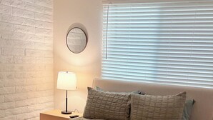 Een slaapkamer met een bed, nachtkastje, lamp, raam met jaloezieën en een bakstenen muur.