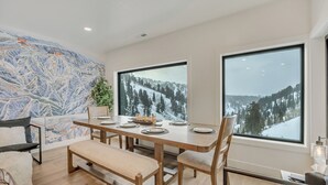 Un área de comedor con un amplio ventanal que ofrece vistas nevadas a las montañas, un mural con un mapa en la pared y una mesa de madera dispuesta para cuatro comensales.