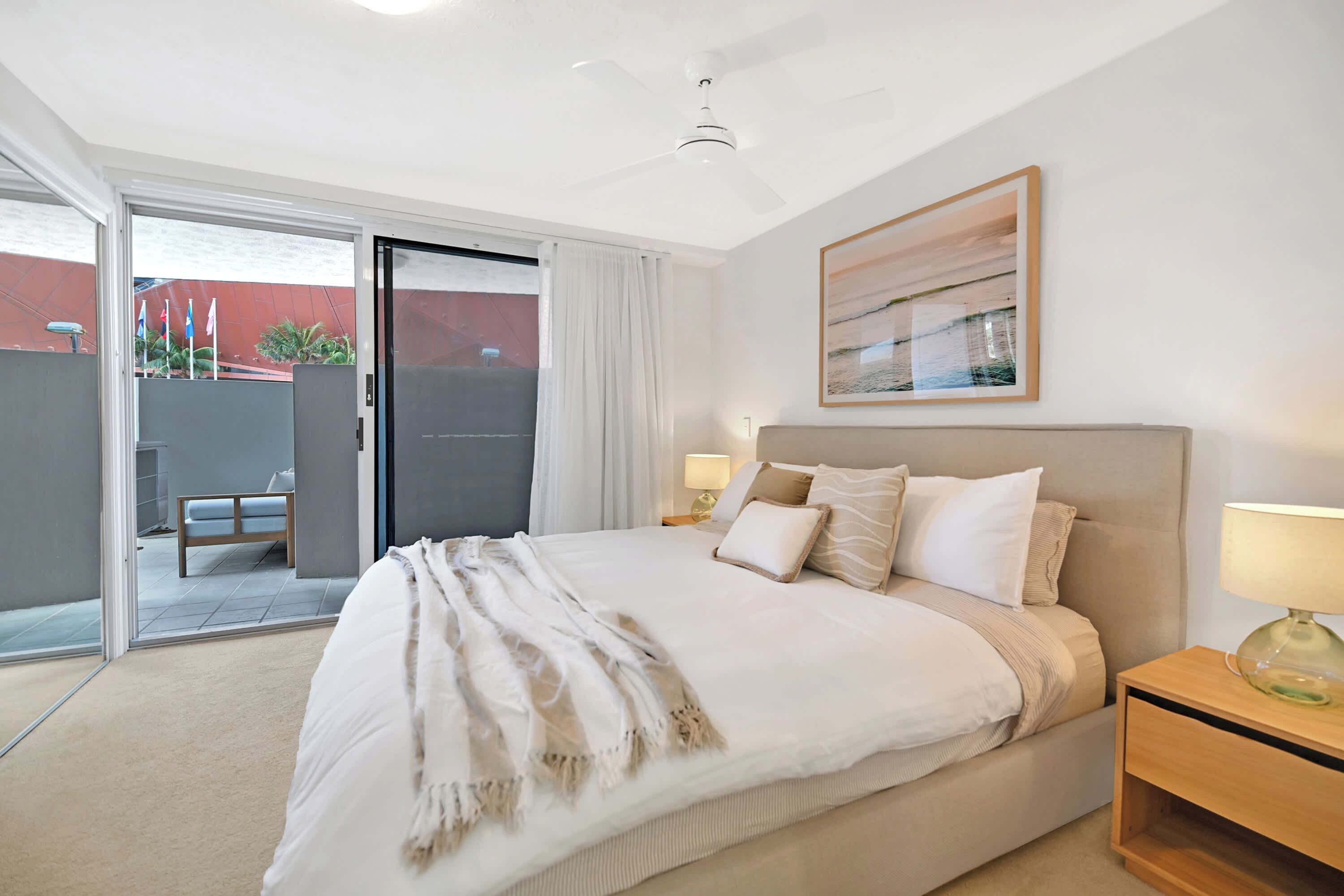 Une chambre à coucher comprenant un lit, une table de chevet, une lampe, un ventilateur de plafond et une porte-fenêtre coulissante donnant sur un espace extérieur.