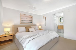 A bedroom with a bed, bedside lamps, a ceiling fan, a framed picture, and a bathroom with a mirror and sink. - Seaside Getaway Large Private Courtyard - Parking (Surfers Paradise)