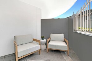 Ein kleiner, abgeschlossener Balkon mit zwei Holzstühlen und einem runden Tisch, mit einer grauen Wand und einem weißen Geländer.
