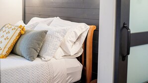 A bed with white bedding and a wooden headboard, a nightstand with a lamp, and a door with frosted glass panels. - One Eyed Odie's Cozy 3-bed, 1-bath home with a relaxing hot tub. (Cove)