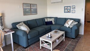 Ein Wohnzimmer mit einem blauen Ecksofa, einem weißen Couchtisch, einem gemusterten Teppich und einem Deckenventilator.