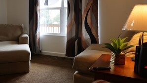 A hotel room with a sofa, a small table with a lamp and a plant, a window with curtains, and a remote control on the table.