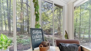 Ein gemütliches Zimmer mit großem Fenster und Blick auf einen Wald, einem Willkommensschild, einem Korbwesen mit Kiefernzapfen und einer gepolsterten Bank.