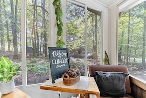 A cozy room with a large window overlooking a forest, a welcome sign, a wicker basket with pinecones, and a cushioned bench.