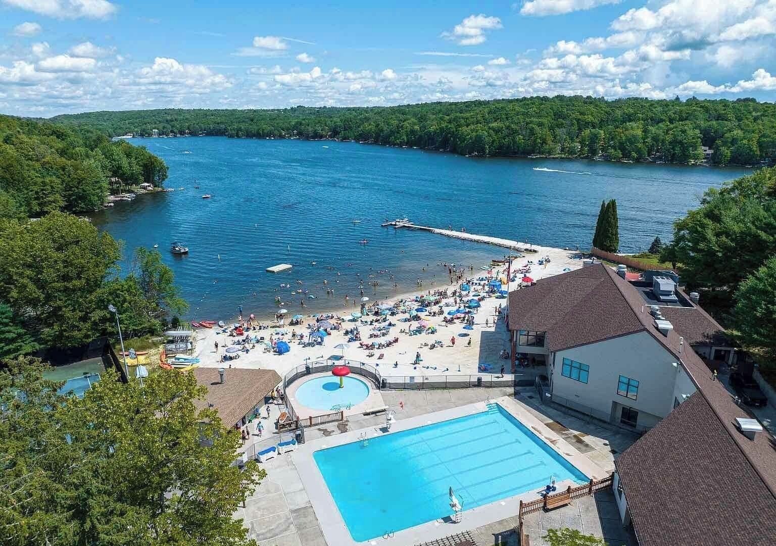 A resort with a swimming pool, beach area, and a dock extending into a lake surrounded by forest.