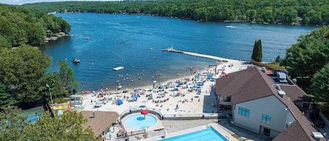A resort with a swimming pool, beach area, and a dock extending into a lake surrounded by forest.