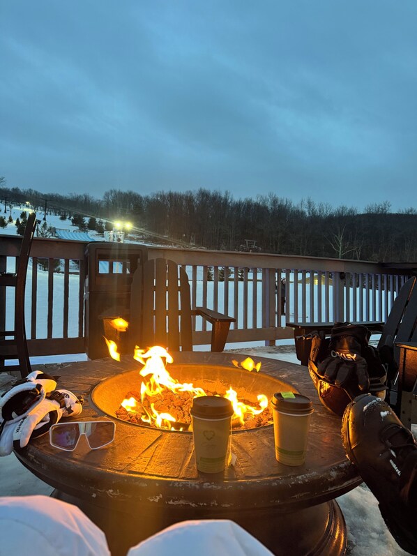 A fire pit table with drinks and gloves, surrounded by a wooden deck with a snowy landscape in the background.