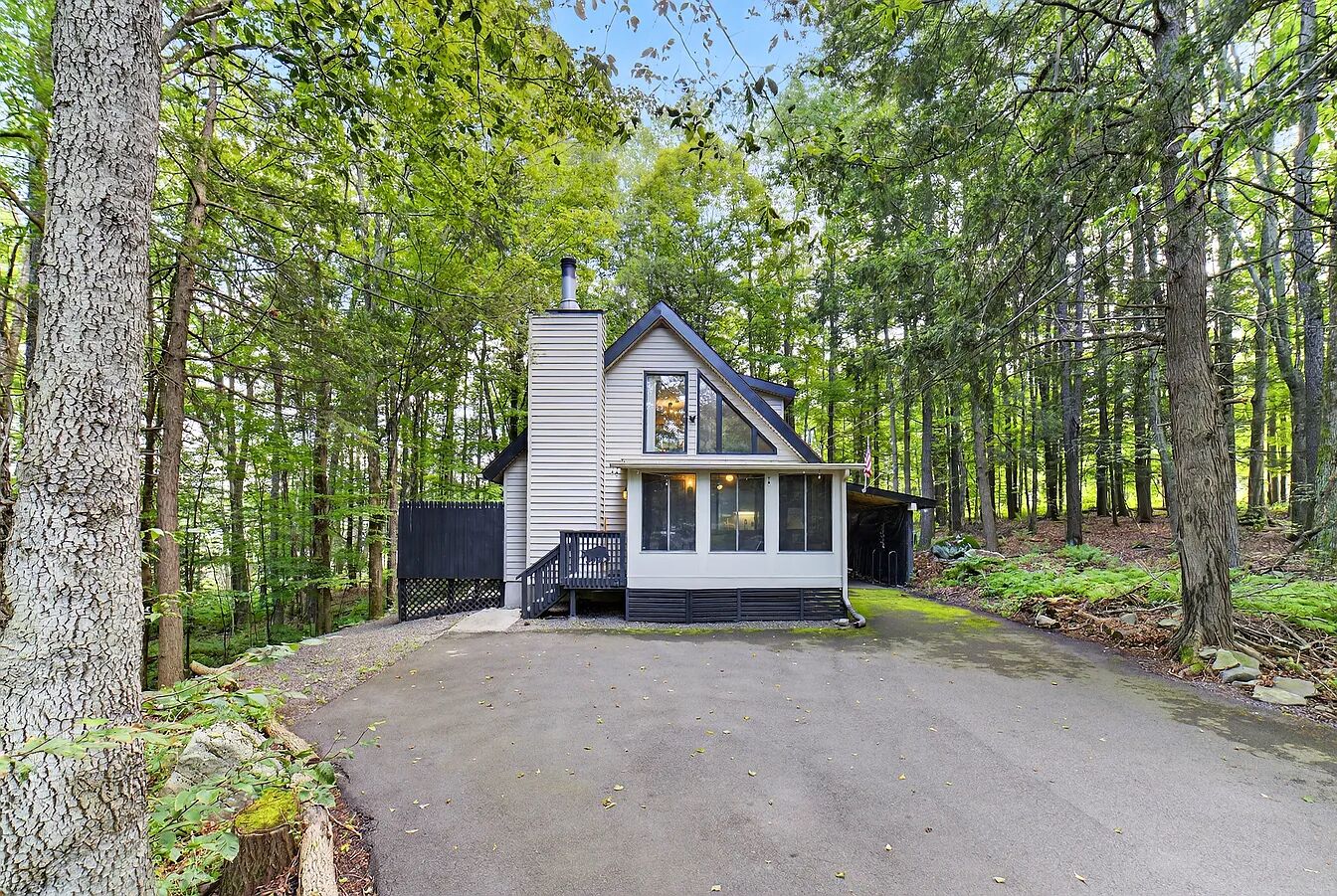 Une cabane dotée d’un porche couvert, d’une cheminée et d’une allée pavée, le tout entouré d’arbres.