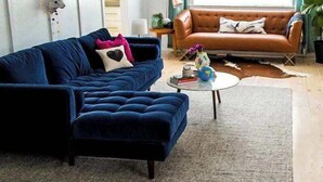 Un salon avec un canapé bleu, un canapé marron, une table basse ronde et une grande fenêtre avec des rideaux.