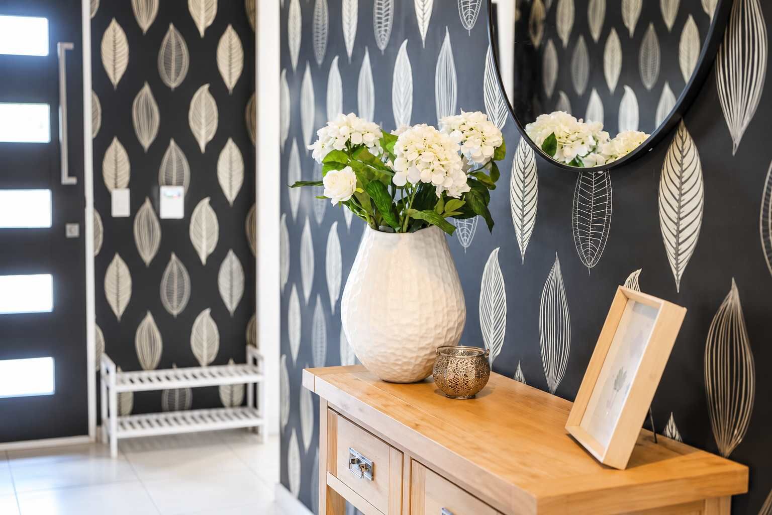 Een kamer met behang in zwart-wit met een bladmotief, een witte vaas met bloemen, een klein glazen potje en een houten bijzettafeltje met een fotolijstje.