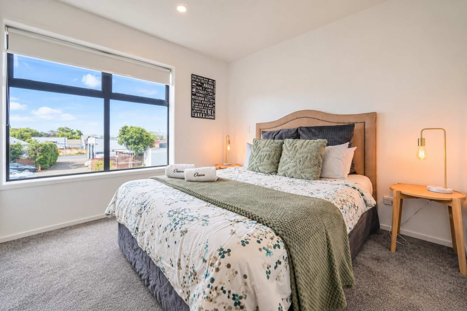 A bedroom with a large window, a bed with a headboard, two bedside tables, and a wall art piece.