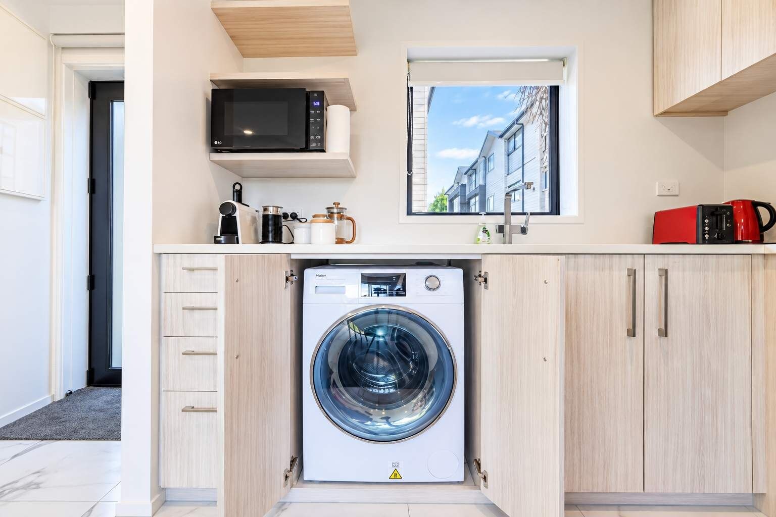 Une cuisine compacte équipée d’un lave-linge, d’un four à micro-ondes et d’une fenêtre donnant sur des immeubles.